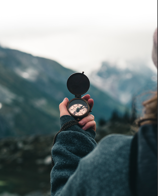 A person holding an open compass, facing cloudy mountain peaks
